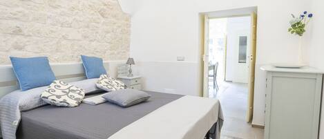 Un dormitorio con pared de piedra, una cama con almohadas estampadas, una mesita de noche con una lámpara y una puerta que da a otra habitación.