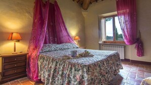 A bedroom with a four-poster bed, floral bedding, a bedside table with a lamp, and a window with curtains.