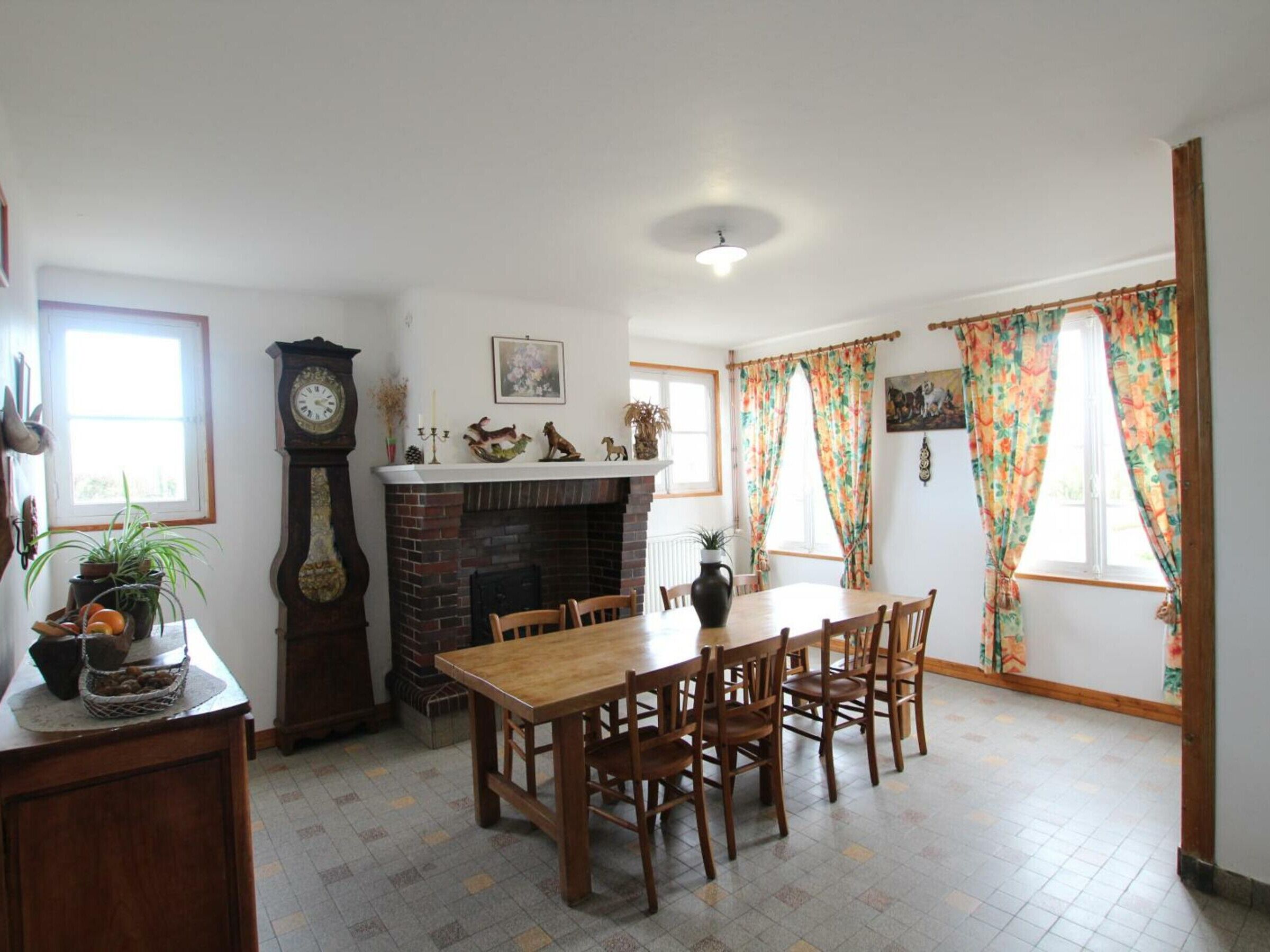 Comedor con chimenea de ladrillo, mesa y sillas de madera. Hay un reloj en la pared y una ventana con cortinas.