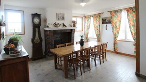 A dining room with a brick fireplace, wooden dining table, and chairs. There is a clock on the wall and a window with curtains. - Charming farm with garden and BBQ in Bérigny (Bérigny)