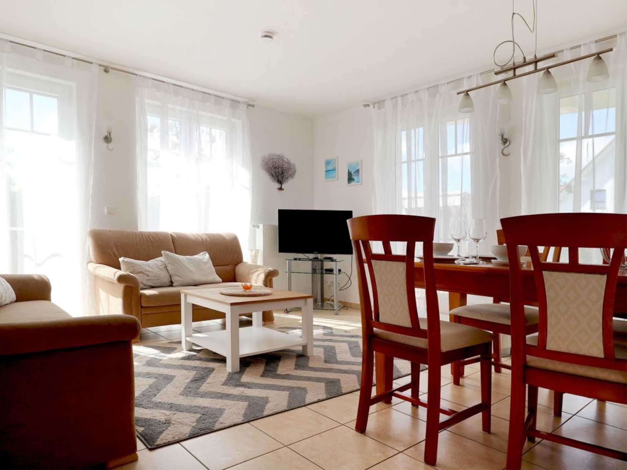 A living room with a sofa, a coffee table, and two wooden chairs. A flat-screen TV is mounted on the wall.
