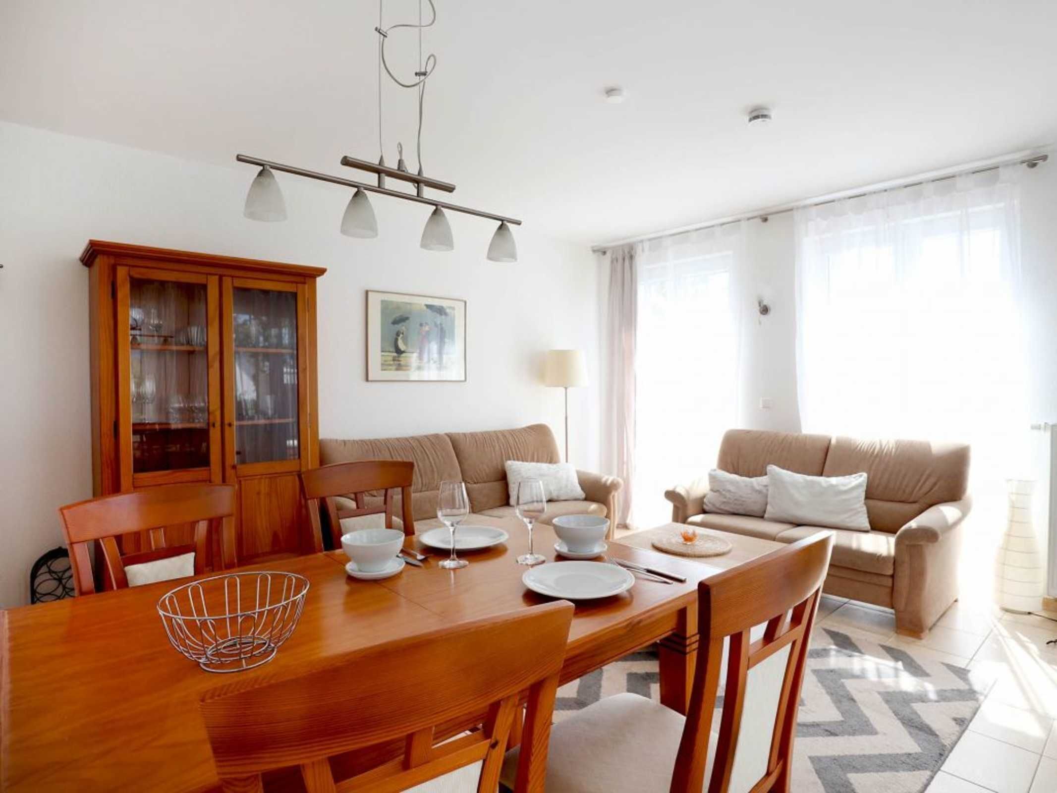 Un coin repas avec une table en bois dressée pour quatre personnes, un meuble en bois doté de portes en verre, un canapé beige et une grande fenêtre ornée de rideaux légers.