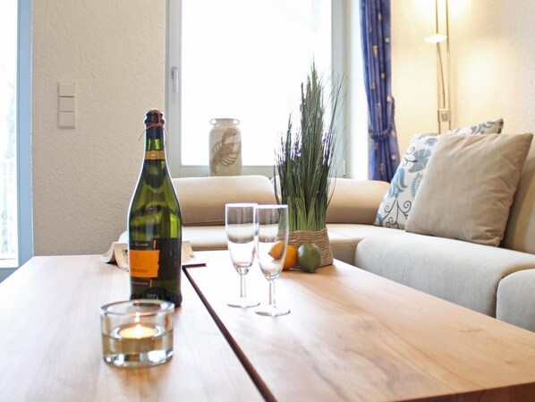 Een kamer met een houten tafel, een fles, twee glaasjes, een kaars, een vaas en een plant.