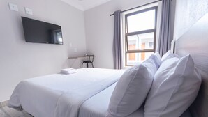 Habitación de hotel con cama, televisor montado en la pared, ventana con cortinas y una pequeña mesa con una silla.
