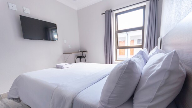 Habitación de hotel con cama, televisor montado en la pared, ventana con cortinas y una pequeña mesa con una silla.