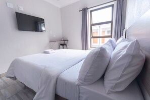 A hotel room with a bed, a TV mounted on the wall, a window with curtains, and a small table with a chair. - Ecomotel Marlboro (Sandton)