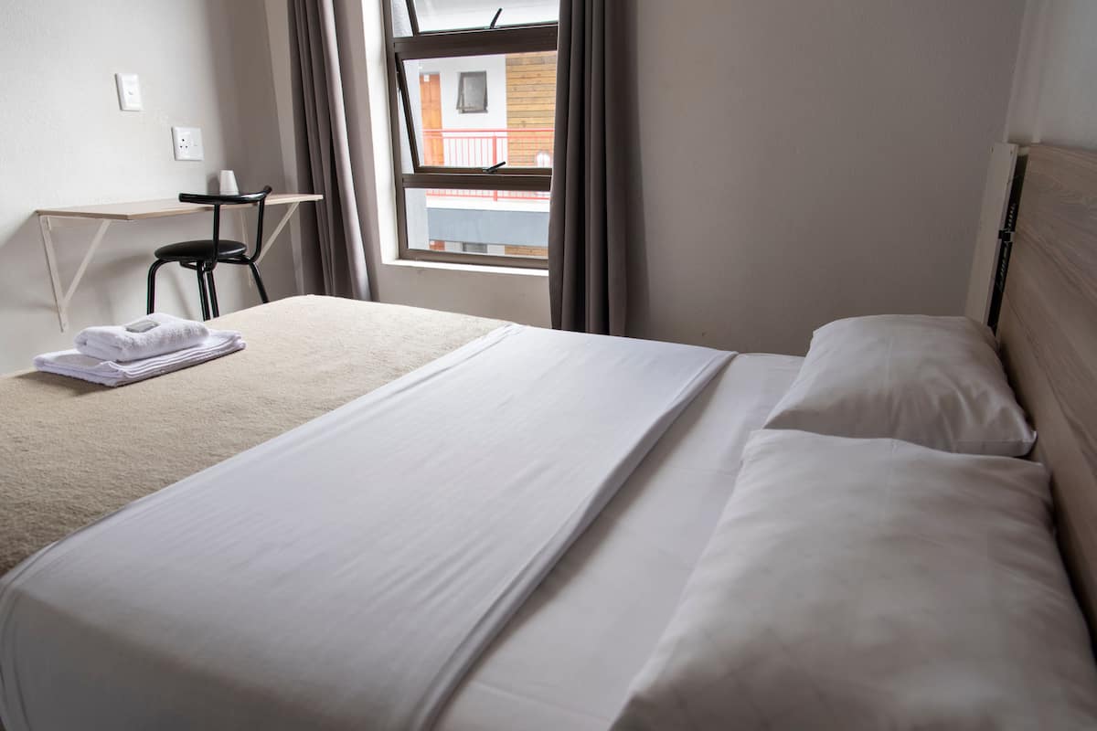 A neatly made bed with white linens, a small desk with a chair, and a window with curtains.