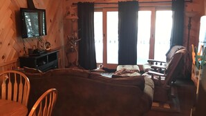 L’intérieur d’un chalet accueillant, avec un plafond en bois, un ventilateur de plafond, un téléviseur, un canapé, une table à manger avec des chaises et une fenêtre avec des rideaux.