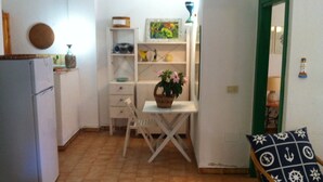 A compact kitchen with a dining area, a white refrigerator, a small table with a basket of flowers, and a chair with a nautical-themed cushion.