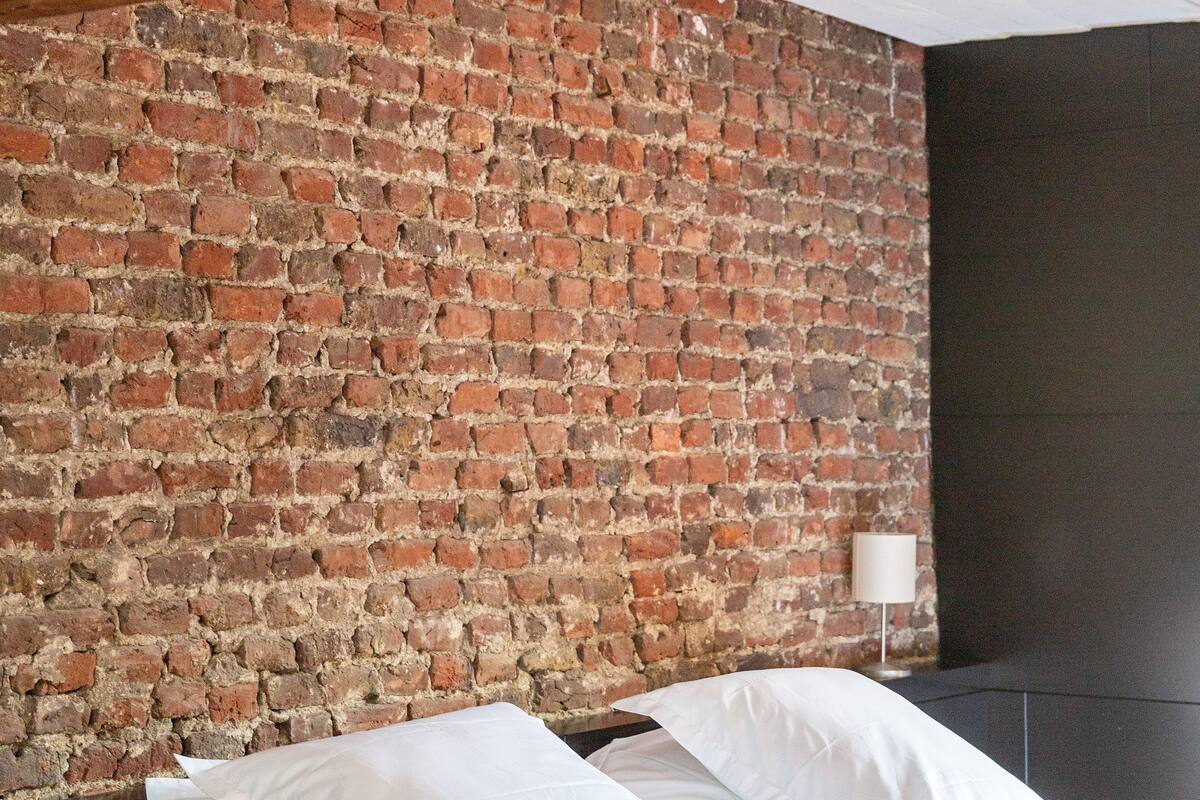 A bedroom with a brick wall, a bed with white linens, and a bedside table with a lamp.