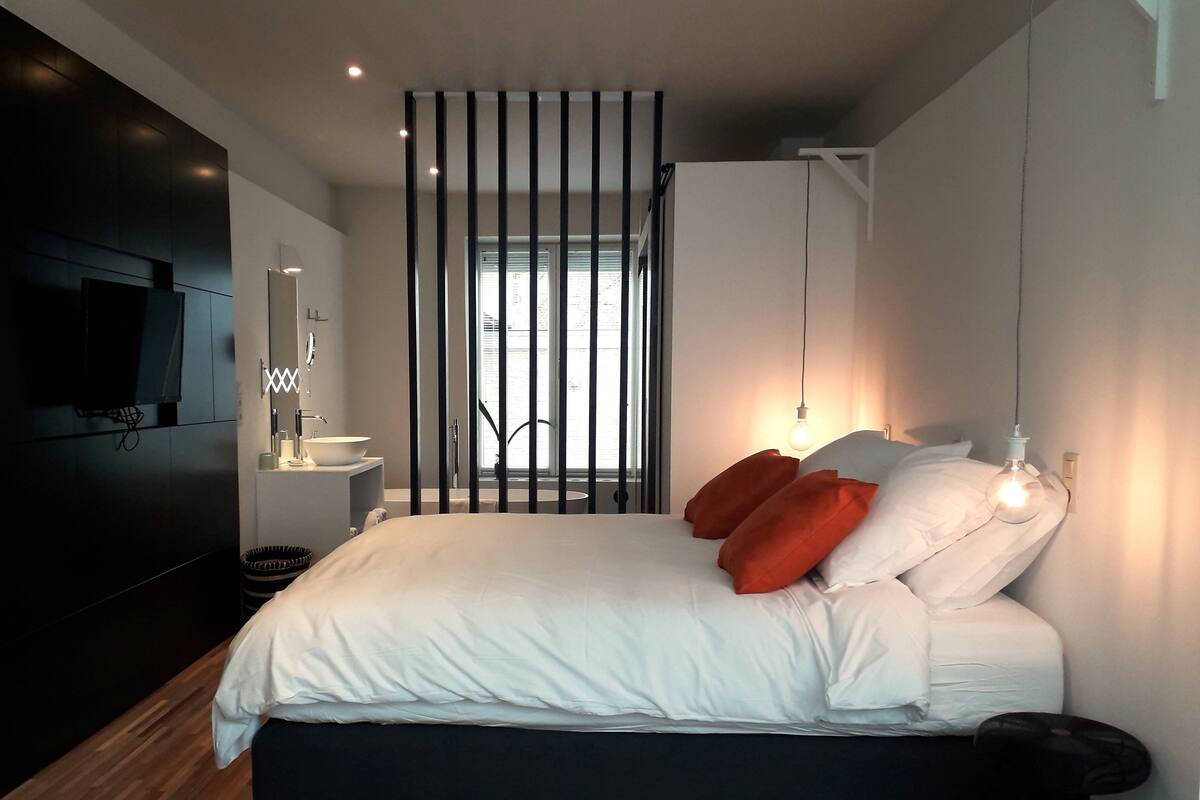 A modern bedroom with a large bed, a wooden floor, a TV, a bedside table, and a window with blinds.
