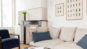 Una sala de estar moderna con un sofá color beige, un sillón azul y una mesa de centro redonda negra. En la pared hay cuadros con imágenes de insectos y una planta pequeña sobre la repisa de la chimenea.