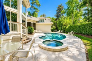 A backyard with a swimming pool, a covered patio with chairs, and a house with a covered deck.