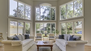 A spacious living room with two white sofas, a wooden coffee table, and large windows offering a view of the outdoors. - 13 Duck Hawk -Relax on the Front porch w/ rockers and a private pool! (Hilton Head Island)