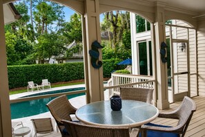A covered outdoor area with a pool, wicker furniture, and a view of the surrounding greenery.