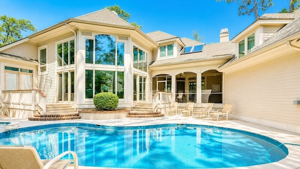 A swimming pool with lounge chairs, a covered patio, and a large house with multiple windows. - 13 Duck Hawk -Relax on the Front porch w/ rockers and a private pool! (Hilton Head Island)