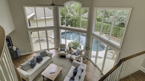 A spacious living area with a large window overlooking a pool and garden, a ceiling fan, and a staircase leading to an upper level. - 13 Duck Hawk -Relax on the Front porch w/ rockers and a private pool! (Hilton Head Island)