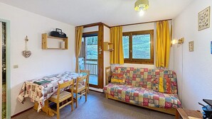 Une pièce chaleureuse avec une table et des chaises en bois, un canapé coloré, une fenêtre avec des rideaux et un luminaire au plafond.