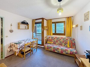 Una habitación acogedora con una mesa y sillas de madera, un sofá colorido, una ventana con cortinas y una lámpara en el techo.