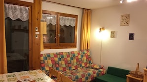 Ein gemütliches Zimmer mit einem Holztisch, einem bunten Sofa und einem grünen Sessel.