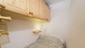 Une petite pièce étroite, avec un lit simple, une armoire en bois au-dessus du lit et une étagère fixée au mur, avec une lampe.