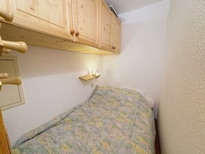 Habitación pequeña y estrecha con una cama individual, un armario de madera sobre la cama y una estantería con luz.