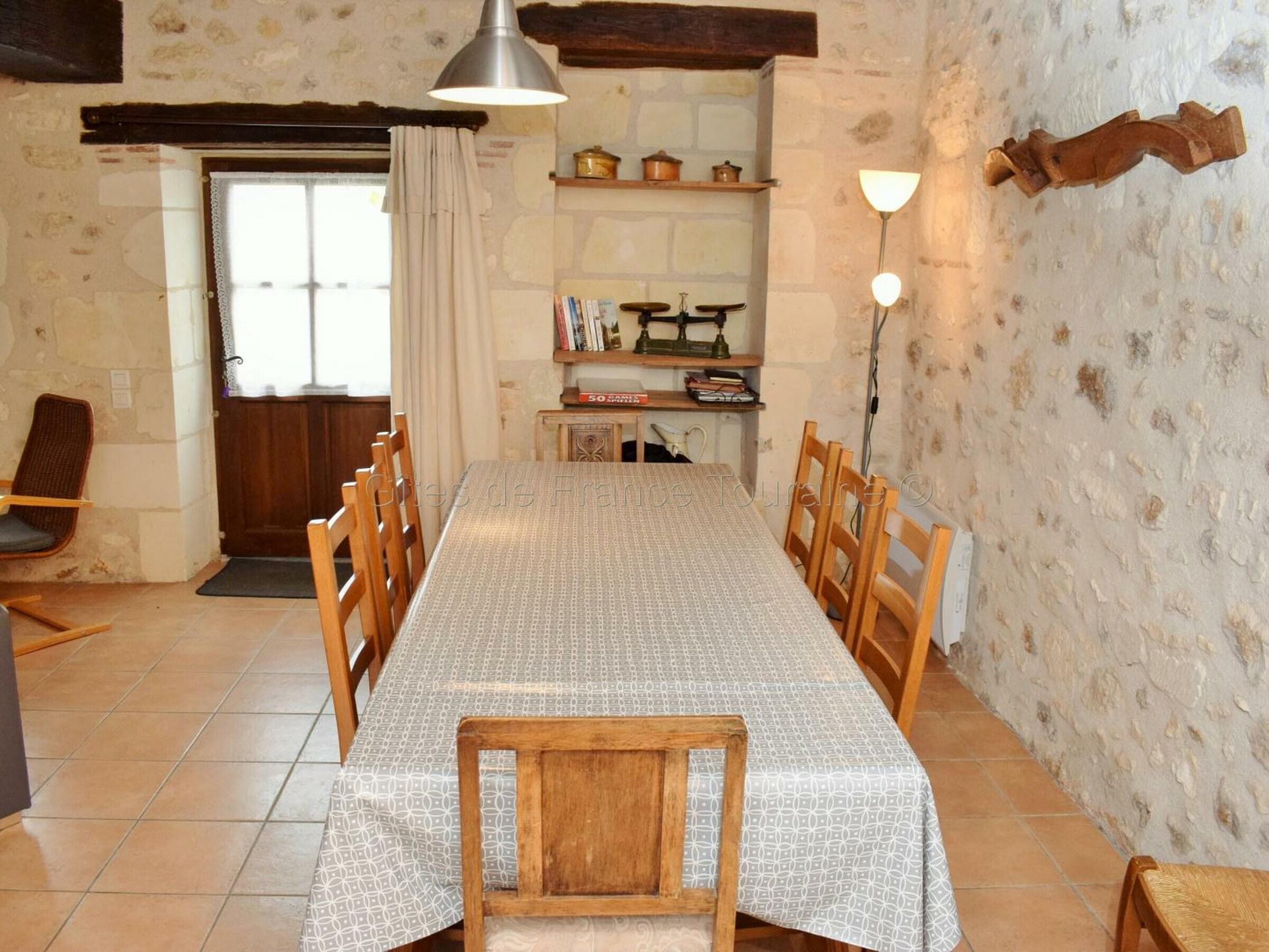 Une salle à manger avec une longue table, des chaises en bois, un lustre et une bibliothèque remplie de livres et d’objets décoratifs.