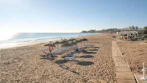 En strand med solstolar, en livboj och en träpromenad som leder till en byggnad med bord och stolar.