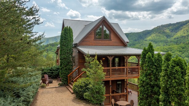A log cabin with a balcony, surrounded by dense forest and hills.