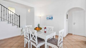 Uma sala de jantar com uma mesa branca, quatro cadeiras, uma cesta de frutas e uma garrafa de vinho.