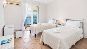 Quarto de hotel com duas camas, uma mesinha, uma cadeira, aparelho de ar condicionado e vista para o mar através de uma varanda.