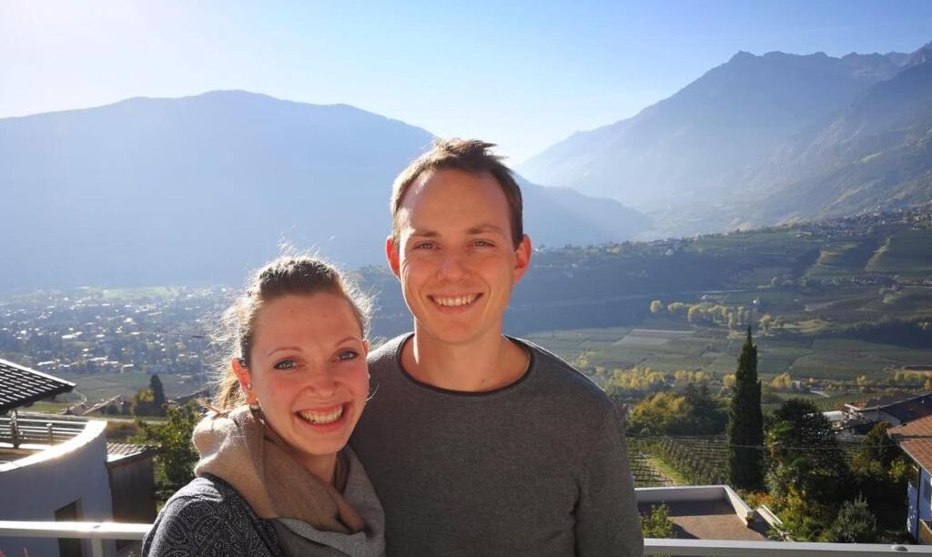 A couple standing on a balcony with a mountainous landscape in the background.