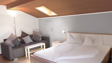 Ein modernes Hotelzimmer mit Holzboden, einem Bett mit weißen Leinen, einem grauen Sofa mit Kissen, einem kleinen weißen Tisch und einem Nachttisch mit Lampe.