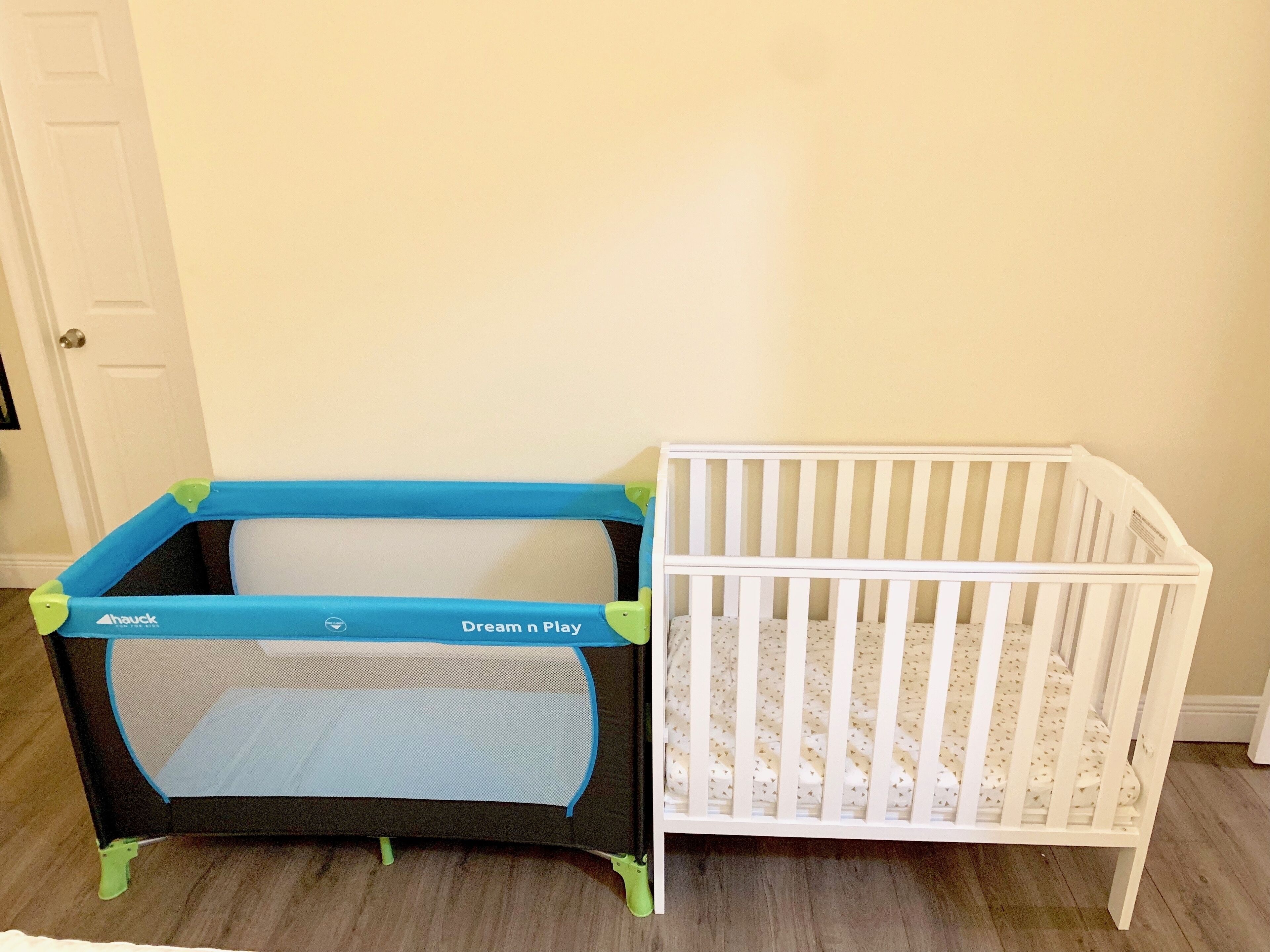 Un parc pour bébé et un berceau dans une pièce dont le sol est recouvert de parquet et qui possède une porte blanche.