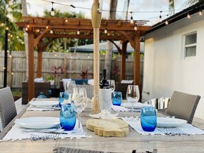 Un área de comedor al aire libre con una pérgola de madera, luces colgantes y una mesa para cuatro personas, con copas de vidrio azul y una botella de vino.