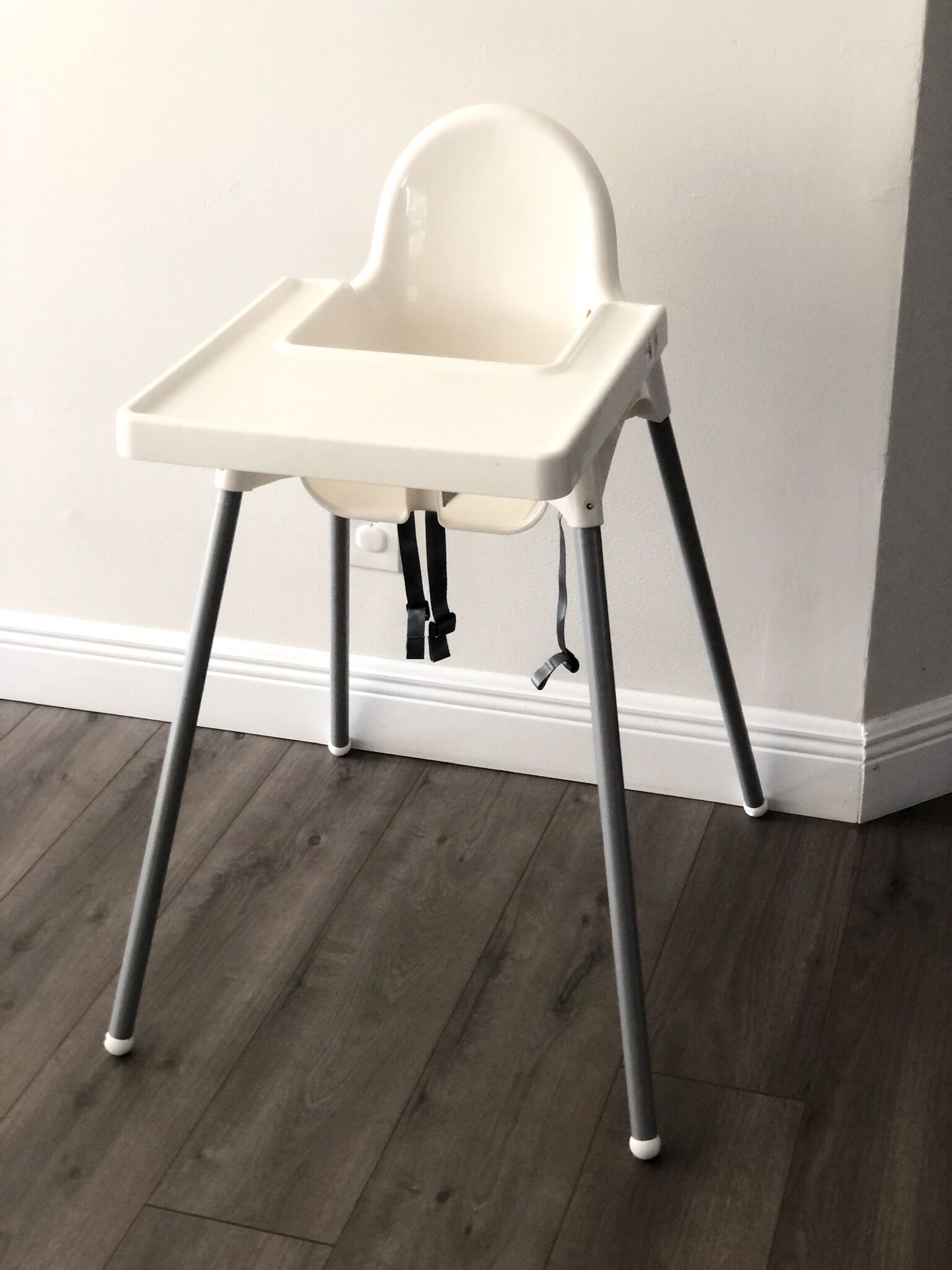 A white high chair with a tray table, positioned against a wall with a wooden floor.