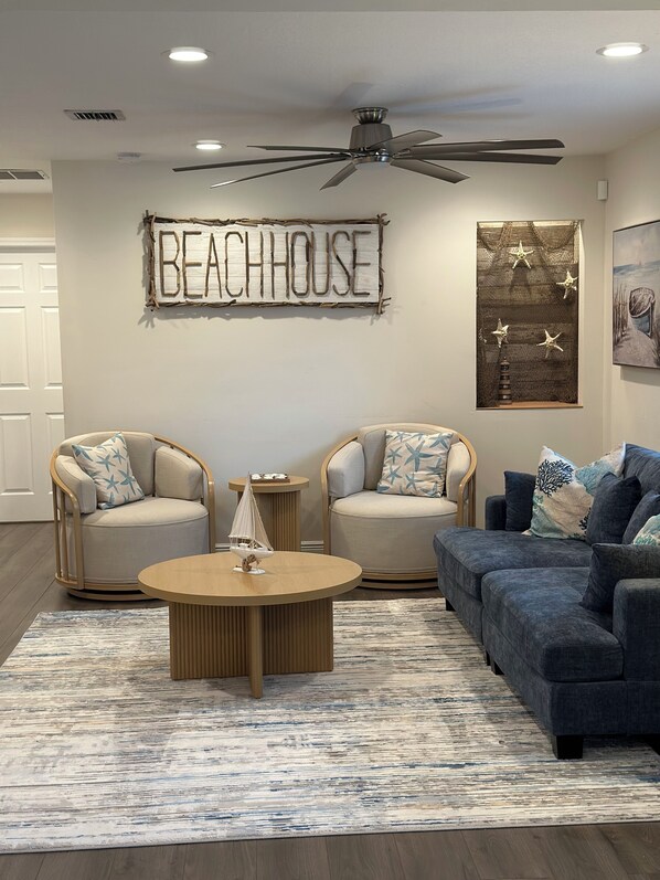 Una sala de estar con dos sillones, un sofá, una mesita de centro y un ventilador de techo. Un cartel en la pared dice "BEACHHOUSE".