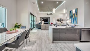 A modern kitchen with a central island, a dining area with a glass table, and a living space with a sofa and TV.