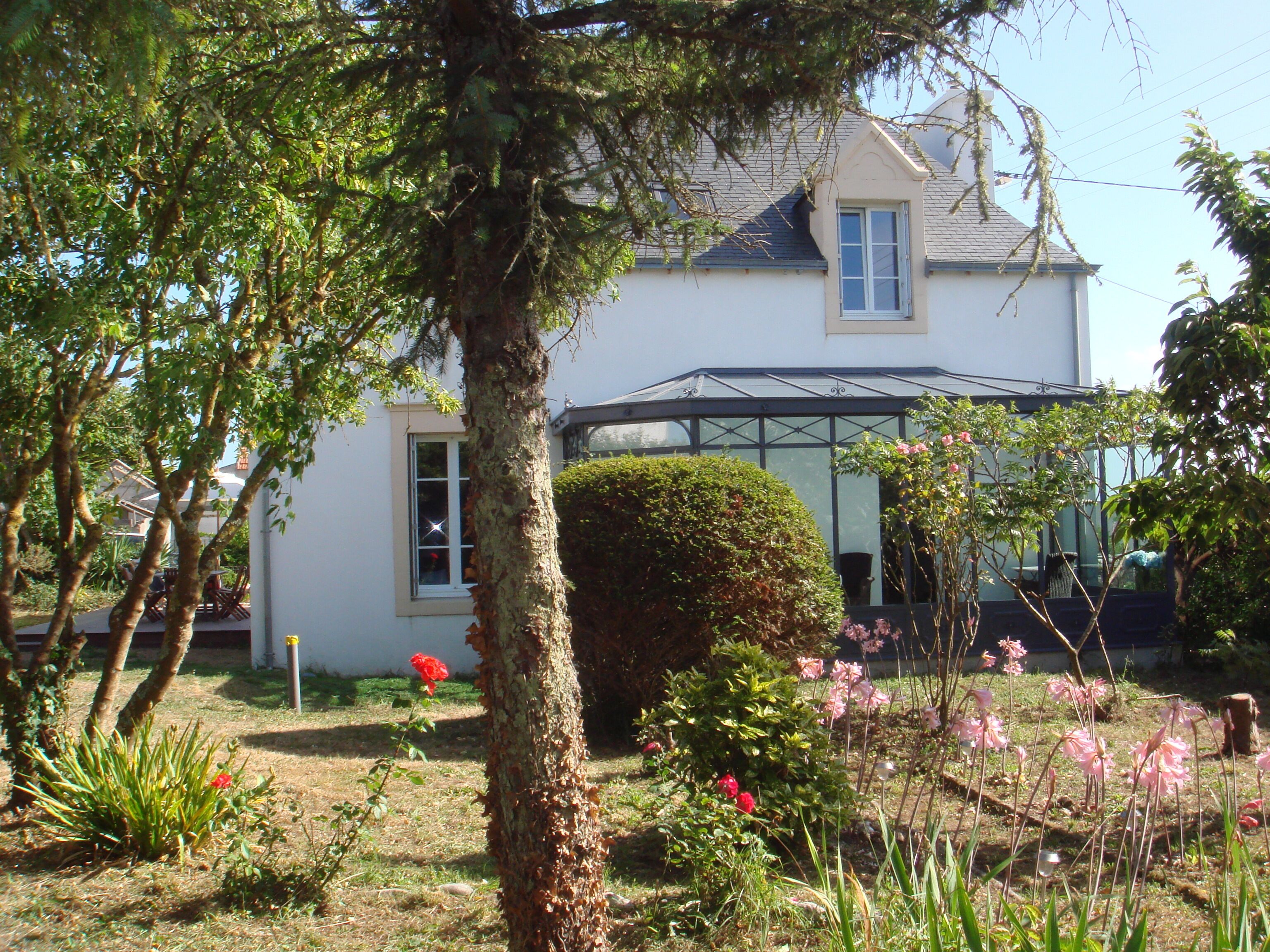 Une maison dotée d’une véranda, entourée d’un jardin où poussent différentes plantes et fleurs.