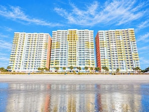 Een hotel met meerdere verdiepingen, balkons, een strand op de voorgrond en heldere lucht.