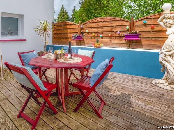 Une terrasse en bois avec une table et des chaises rouges, une statue et un mur bleu agrémenté de panneaux de bois et de décorations colorées.