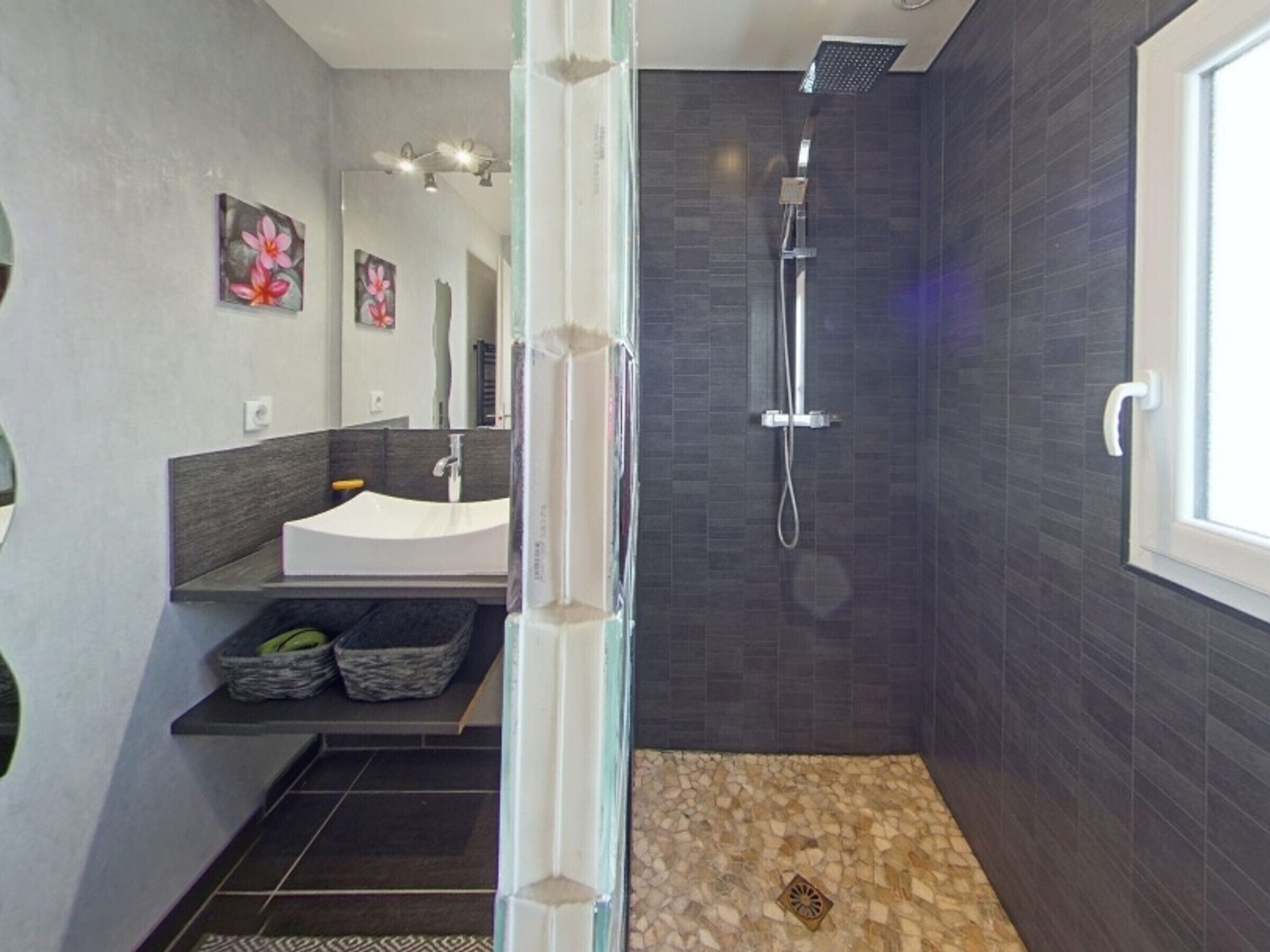 A modern bathroom with a glass shower enclosure, dark tiled walls, a white sink, and decorative flower paintings.