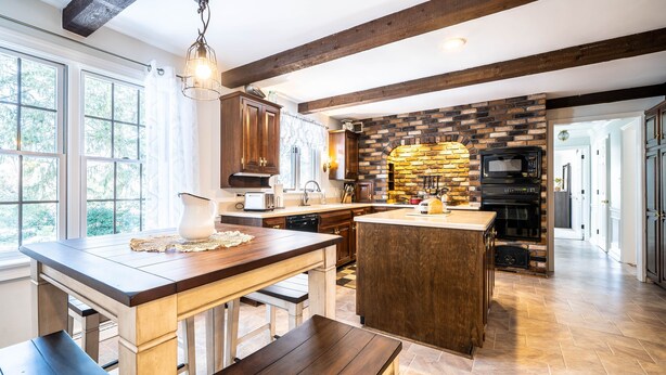 A kitchen with a large island, wooden beams, a brick wall, and modern appliances. - The Evergreen Home In Lancaster County PA. (Bird in Hand)