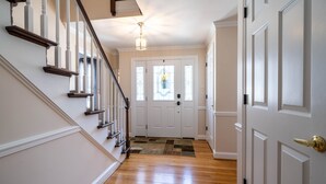 A hallway with wooden flooring, white walls, a staircase with dark railings, and a door with a glass window. - The Evergreen Home In Lancaster County PA. (Bird in Hand)