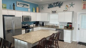 Una cocina con armarios blancos, un refrigerador de acero inoxidable, un microondas y una encimera de granito. Hay tres sillas de madera y una pequeña isla con un fregadero.