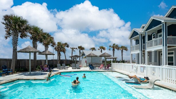 Una piscina con palmeras, sillas de descanso y un edificio con balcones.