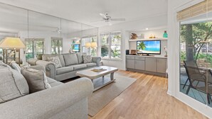 A modern living room with a grey sofa, a coffee table, and a TV. There is a kitchen area with cabinets and a window. - Hidden Dunes First Floor Cottage Steps from the Pool, Rare 1 Bedroom, 2 Bathroom (Miramar Beach)