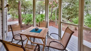 A covered outdoor seating area with a table and chairs, surrounded by glass walls and a view of trees and plants. - Hidden Dunes First Floor Cottage Steps from the Pool, Rare 1 Bedroom, 2 Bathroom (Miramar Beach)