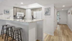A modern kitchen with a marble countertop, white cabinets, and a built-in microwave. There are three stools at the counter. - Hidden Dunes First Floor Cottage Steps from the Pool, Rare 1 Bedroom, 2 Bathroom (Miramar Beach)