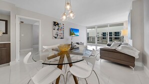 Una sala de estar moderna con una mesa de comedor de vidrio, sillas blancas, un sofá marrón y un amplio ventanal con vistas a la ciudad.